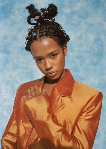 Portrait of a young adult female model wearing a copper-colored blazer with decorated braided hair, set against a blue mottled background.