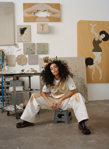 A woman with curly hair poses in an artist's studio, seated on a stool with paintings and art supplies in the background.