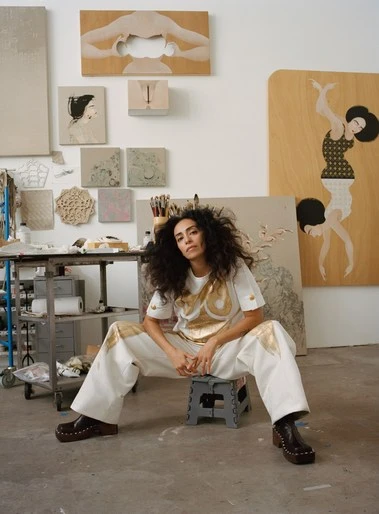 A woman with curly hair poses in an artist's studio, seated on a stool with paintings and art supplies in the background.