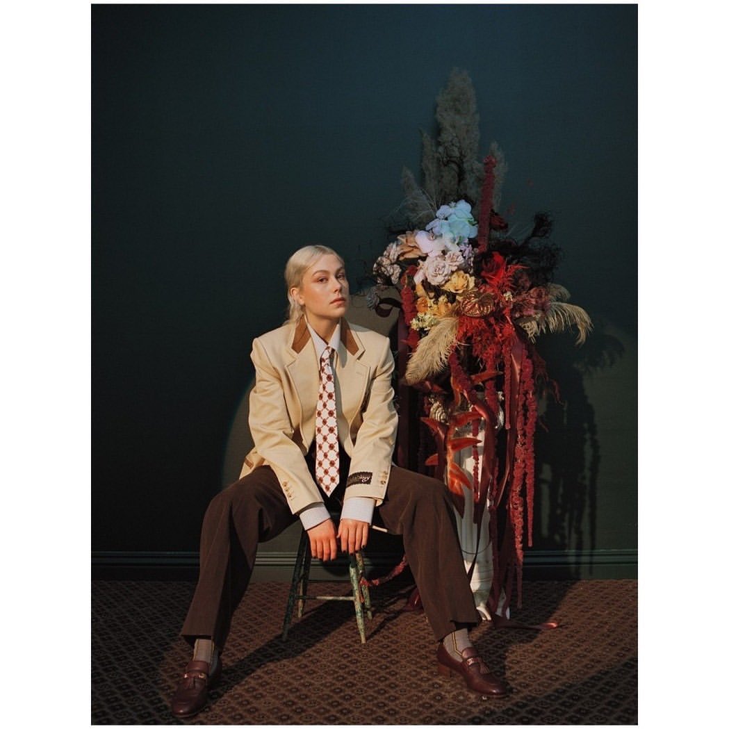 Phoebe Bridgers sits in a beige blazer, brown trousers, and a checkered tie next to a large floral arrangement, indoor setting.