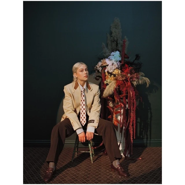 Phoebe Bridgers sits in a beige blazer, brown trousers, and a checkered tie next to a large floral arrangement, indoor setting.