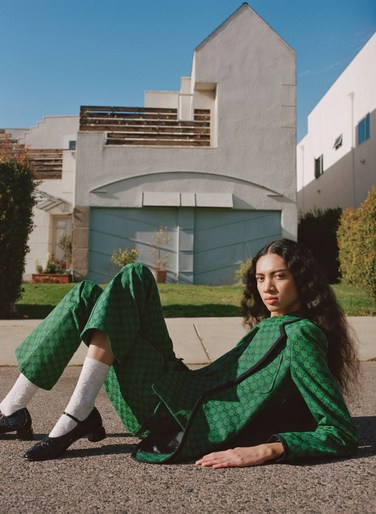 A young woman wearing a green patterned suit and black shoes poses on an asphalt street with white houses and a blue sky in the background.