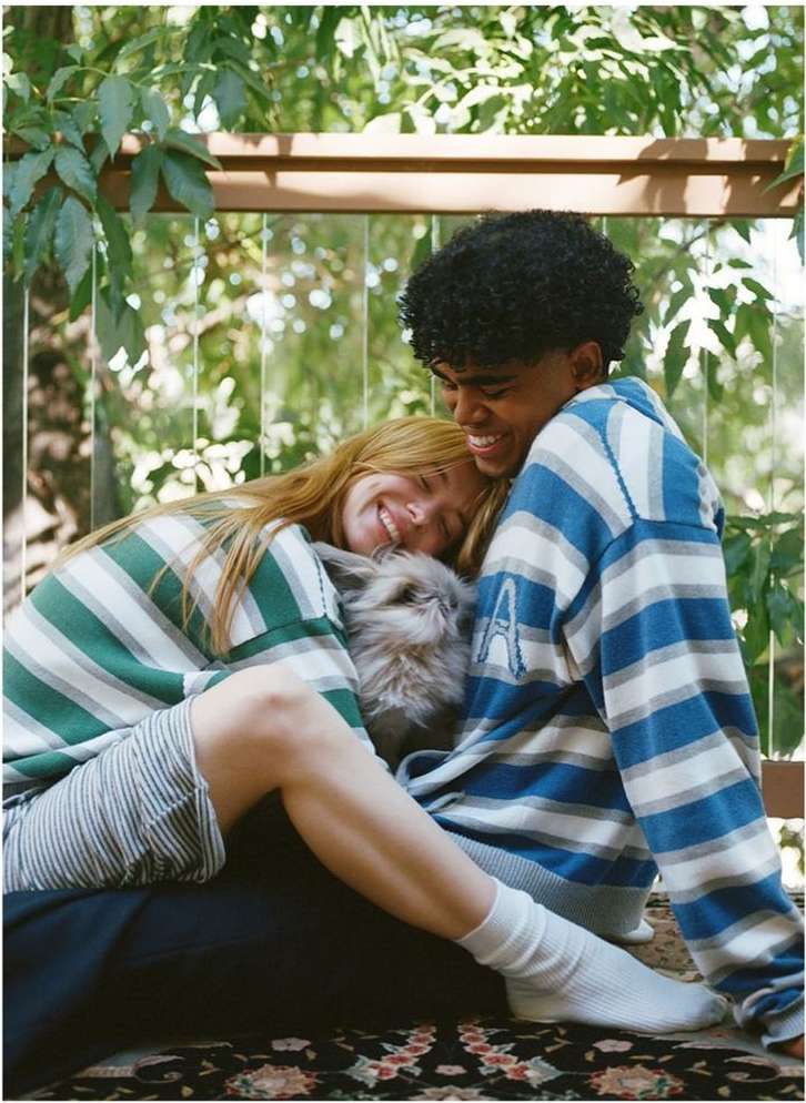 A smiling couple, an adult man and woman, cuddle with a small fluffy dog outdoors, wearing matching striped tops and cozy socks.