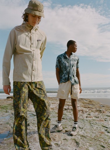 Two adult males model outdoor apparel on a rocky beach with ocean waves and a cloudy sky in the background, captured in natural light.