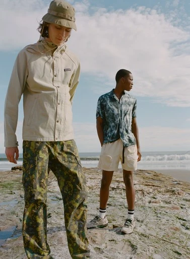 Two adult males model outdoor apparel on a rocky beach with ocean waves and a cloudy sky in the background, captured in natural light.