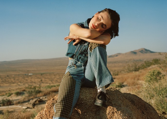 Woman in denim vest and patched jeans seated on a rocky outcrop outdoors in a desert landscape by Daria Kobayashi Ritch