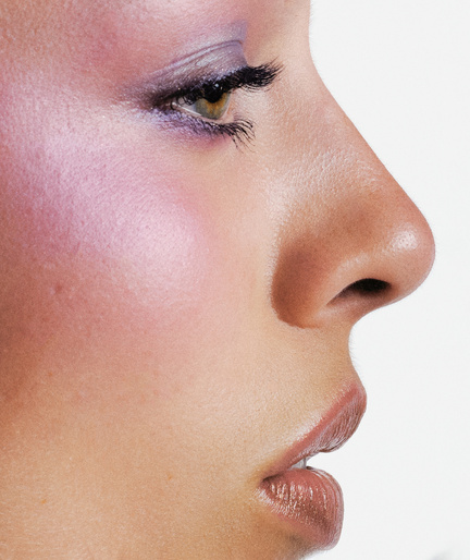Close-up profile portrait of a woman wearing purple eyeshadow and soft pink blush against a plain white background