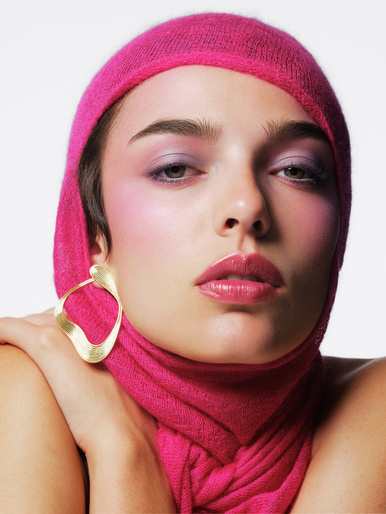 Close-up of a woman with pink eyeshadow and a pink headscarf, wearing a gold earring, posing in front of a white backdrop.