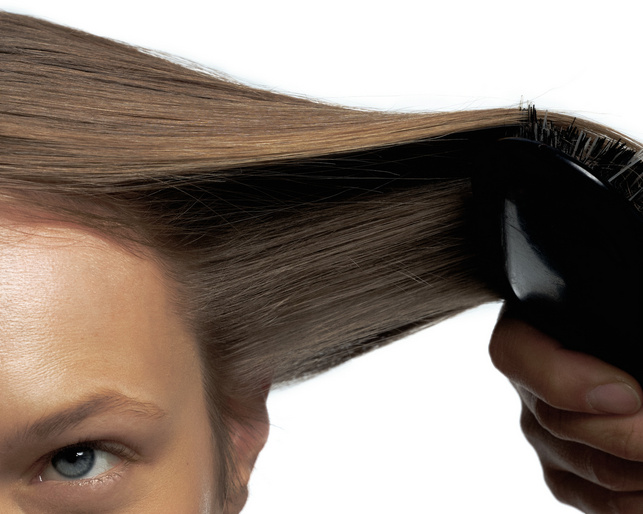 Close-up of an adult female brushing her long, light brown hair with a black hairbrush against a stark white background in a beauty shot.