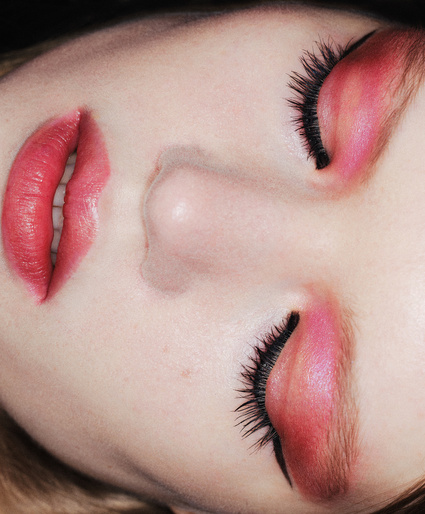 Close-up portrait of an adult female face showcasing glamorous makeup, including bright red lipstick and shimmering pink eyeshadow and long eyelashes.