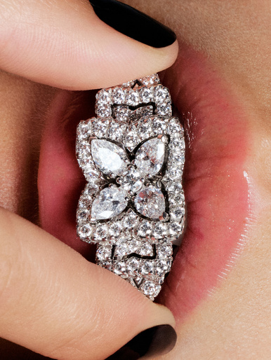 Close-up of a woman's lips with a diamond jewelry piece held between her fingers and mouth, showcasing black nail polish.
