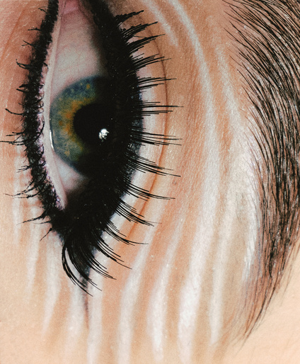 Extreme close-up of a human eye with long lashes, makeup, and visible skin texture illuminated by soft, reflected lighting.
