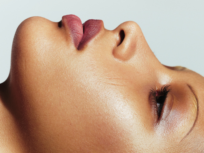 Close-up beauty shot of a light-skinned young adult female's face with shiny skin, pink lips, and closed eye. Grey gradient background.