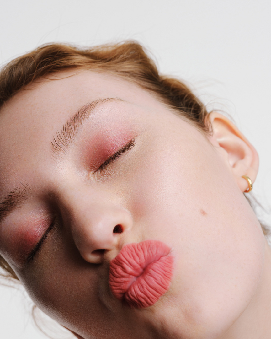 Close-up of a young woman with fair skin and red hair in a beauty shot wearing pink eyeshadow and coral lipstick with pursed lips.