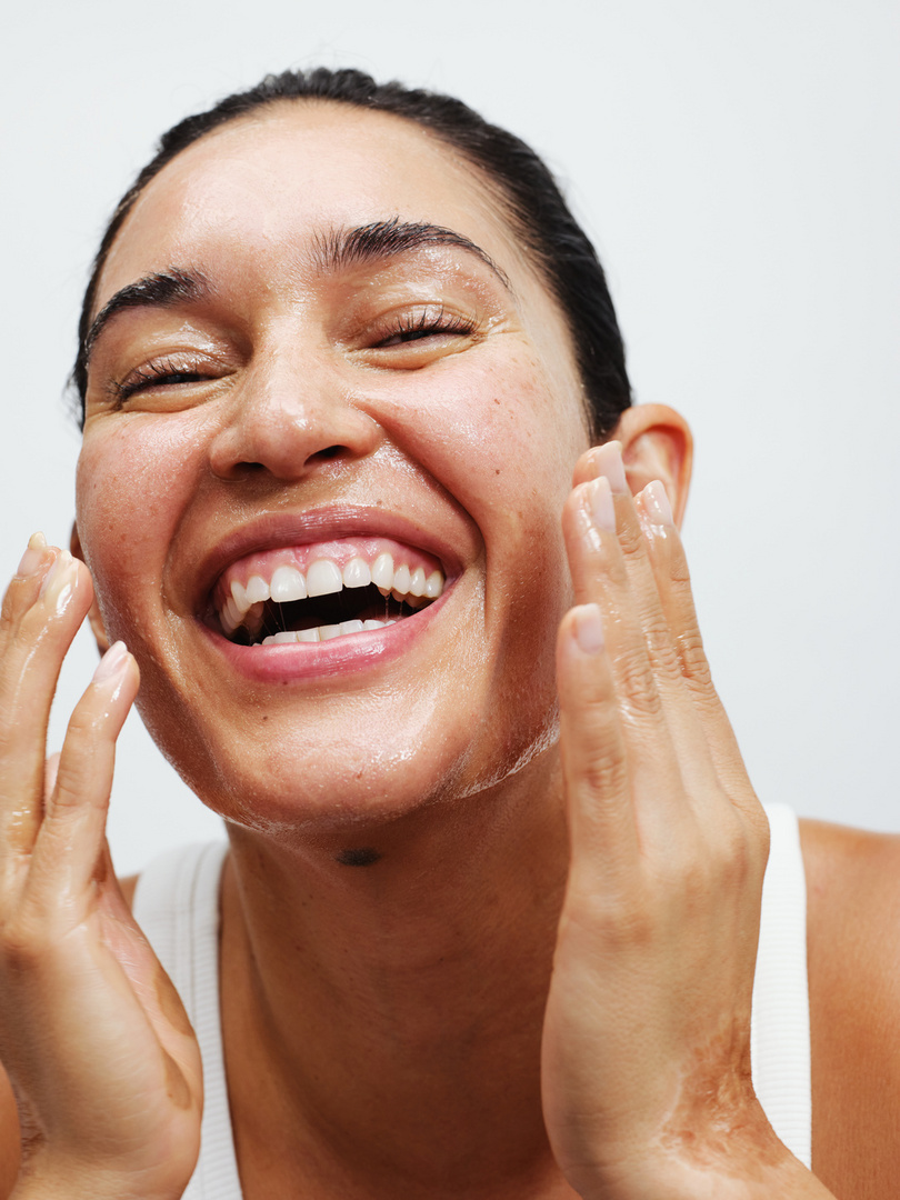 Close-up shot of a smiling adult woman with her eyes closed, applying a clear liquid to her face, set against a plain white background.