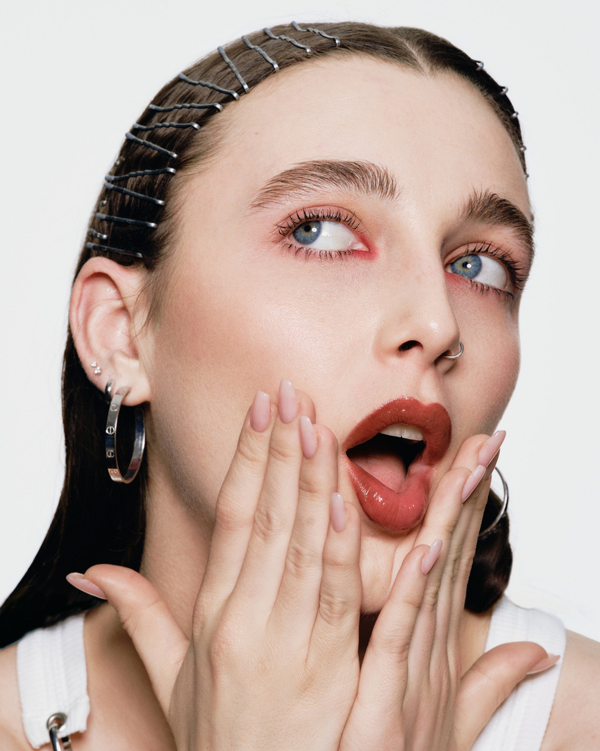 Close-up portrait of a woman with nude nail polish, glossy red lipstick, an open mouth, and a beaded headband against a white background.