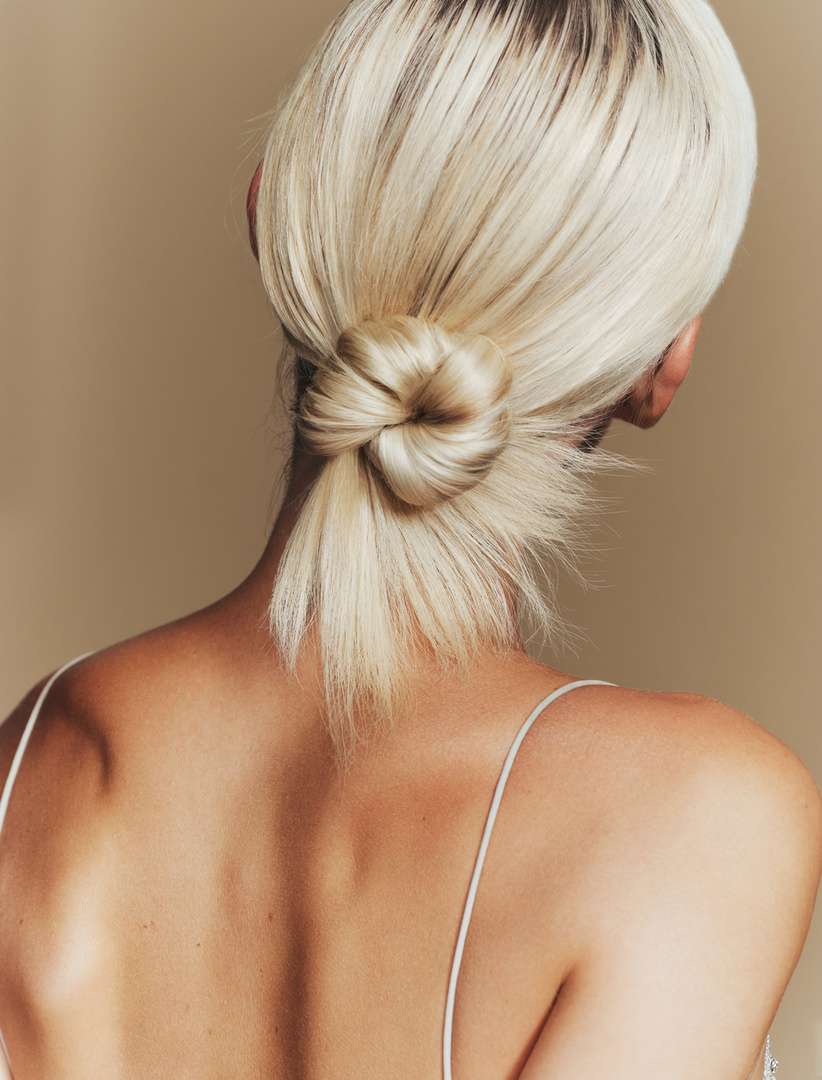 Back view of a light-skinned woman with blonde hair styled in a low knot; she is wearing a spaghetti-strap top against a neutral backdrop.