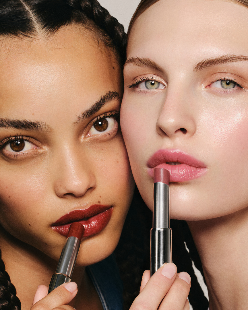 Two women pose closely, each applying lipstick. One model has darker skin and braided hair, the other fair skin and light eyes.