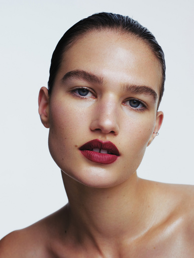 Close-up beauty portrait of a woman with slicked-back dark hair and bold burgundy lipstick against a white background