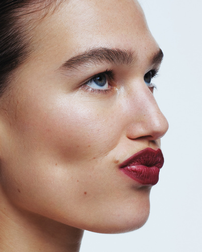 Close-up studio shot of a woman with fair skin, blue eyes, dark red lipstick, and thick eyebrows against a plain white background.