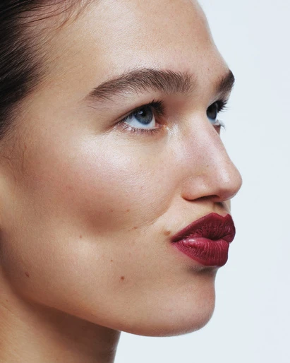Close-up studio shot of a woman with fair skin, blue eyes, dark red lipstick, and thick eyebrows against a plain white background.