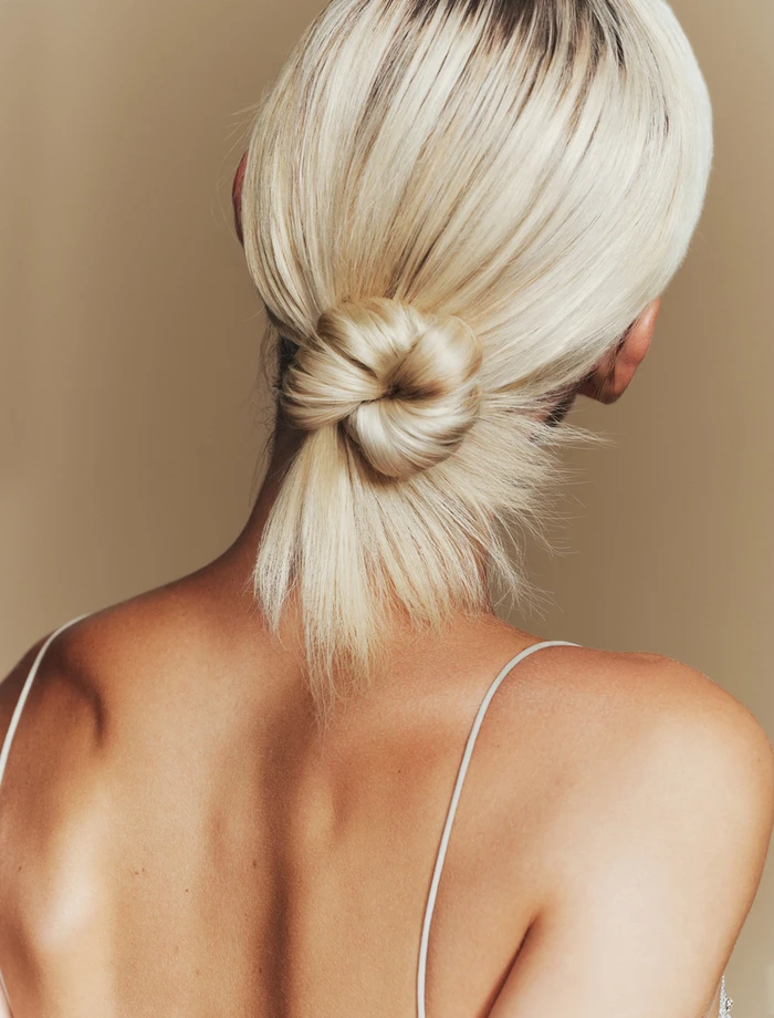 Rear view of a woman with blonde hair styled in a low knot wearing thin straps against a neutral backdrop