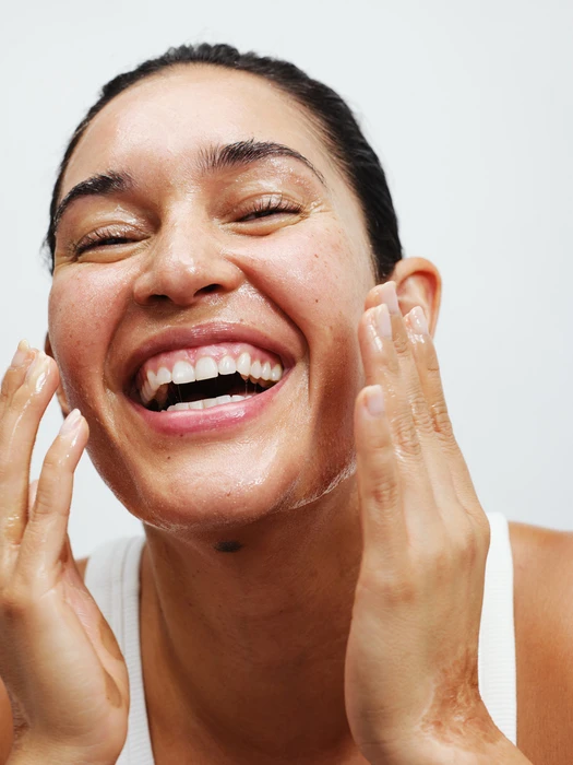 Close-up portrait of a smiling woman with glossy skin holding her hands up near her face against a plain background