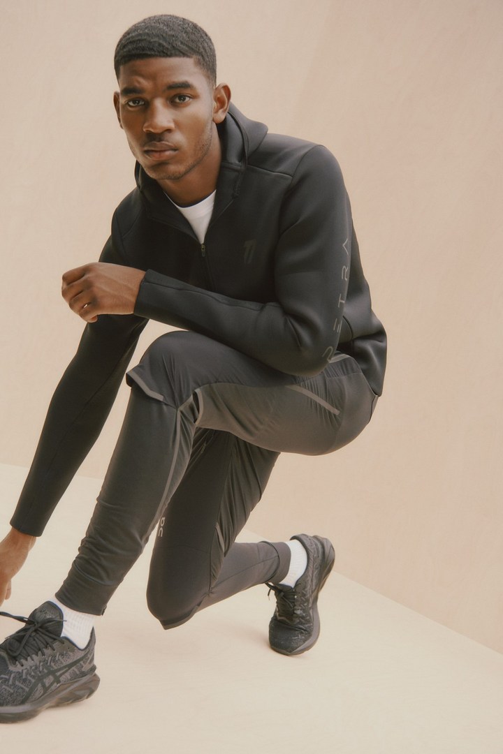 A dark-skinned male model poses in activewear, kneeling indoors with black sneakers against a peachy background in a fashion setting.