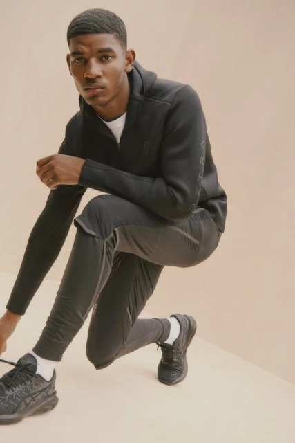 A dark-skinned male model poses in activewear, kneeling indoors with black sneakers against a peachy background in a fashion setting.