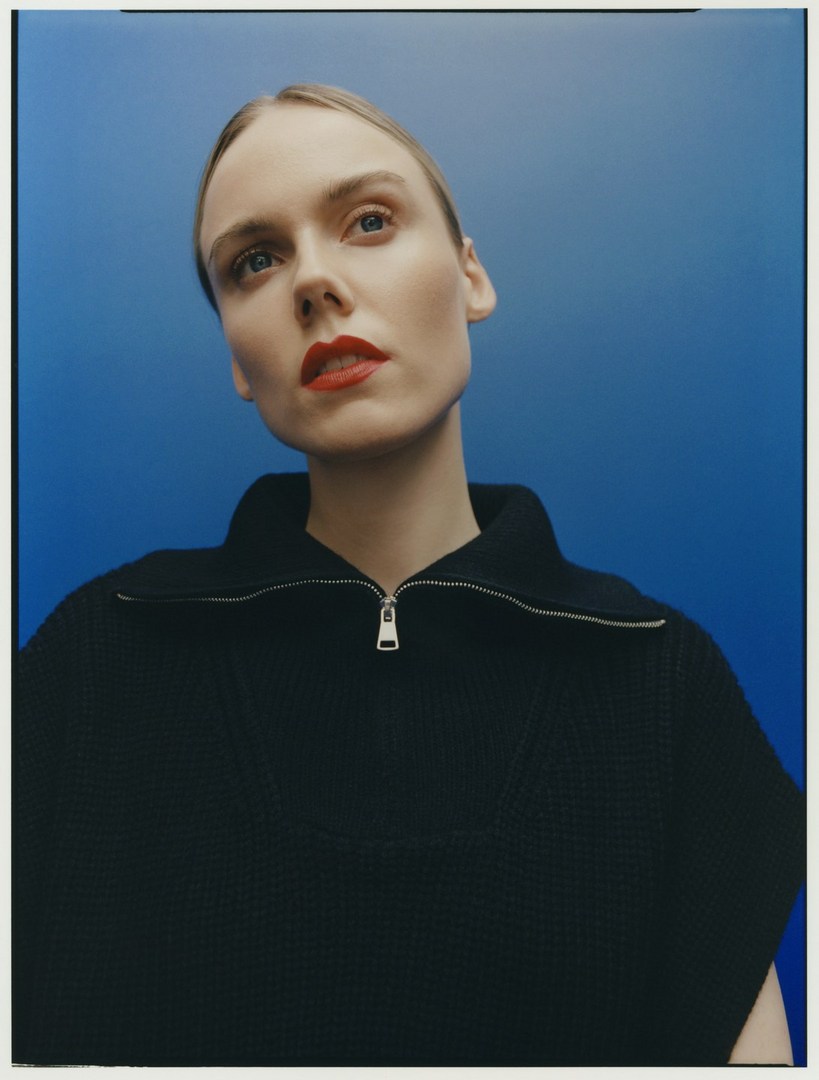 Low-angle shot of a light-skinned woman with red lipstick and a black zip-up sweater against a blue gradient background in a studio setting.