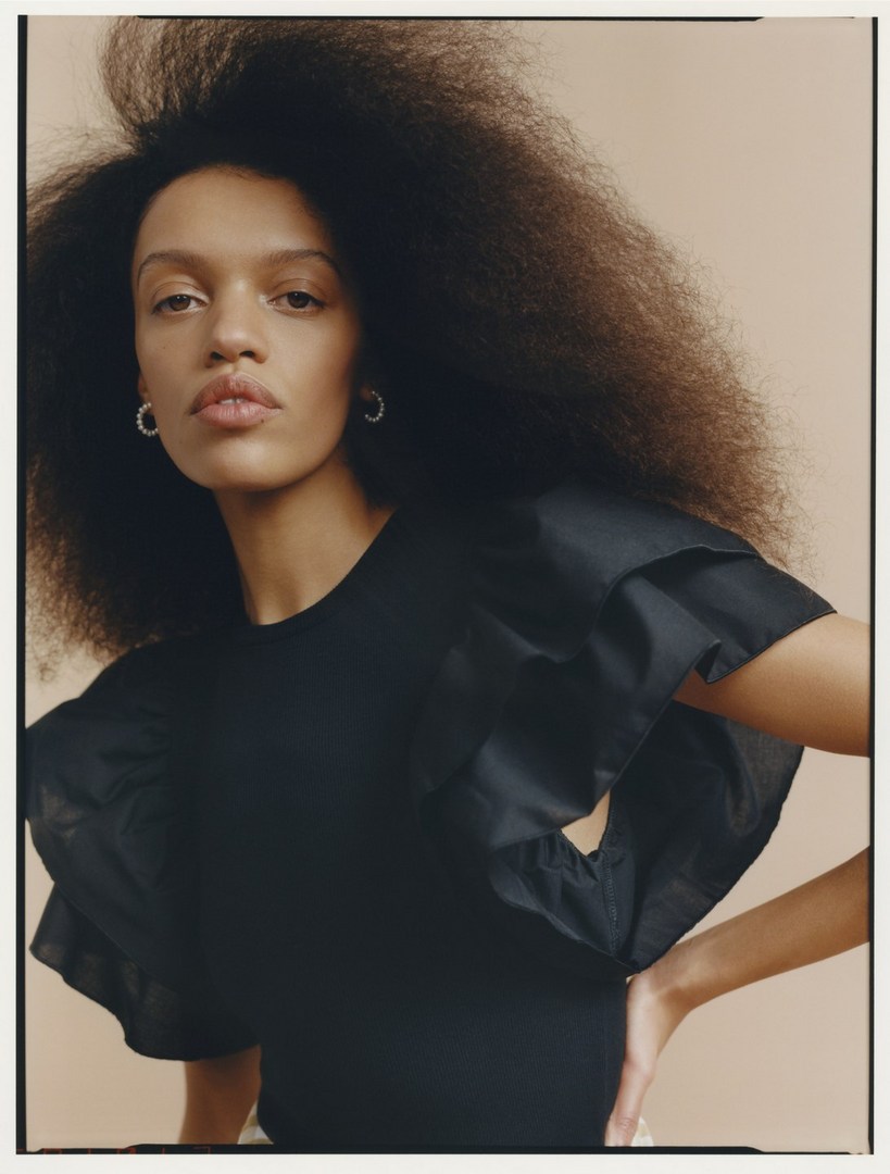 Portrait of a woman with voluminous dark hair wearing a black ruffled top and small silver hoop earrings against a beige backdrop.