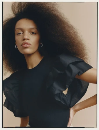 Portrait of a woman with voluminous dark hair wearing a black ruffled top and small silver hoop earrings against a beige backdrop.