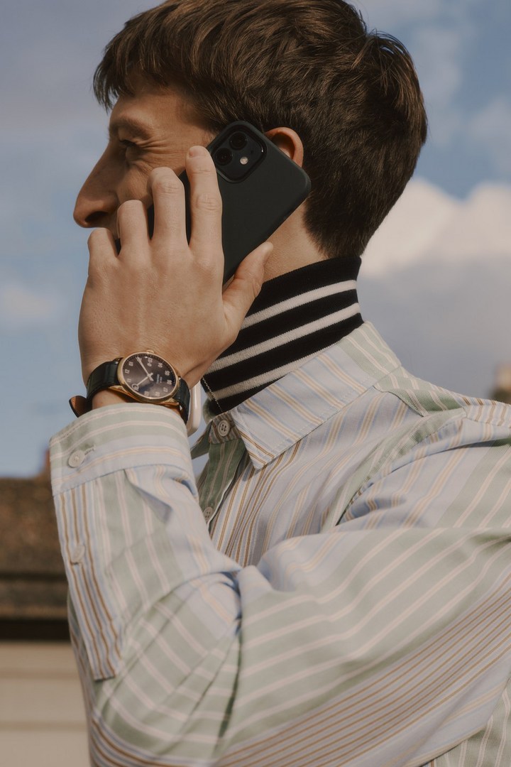 Man talking on smartphone outside with light striped shirt, black and white striped turtleneck, gold watch, blue skies, and sunlight.