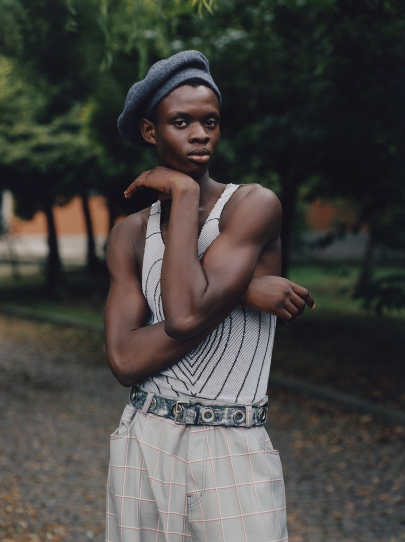 Young adult with a beret posing outdoors. The subject has dark skin and is wearing a sleeveless top and checked trousers.