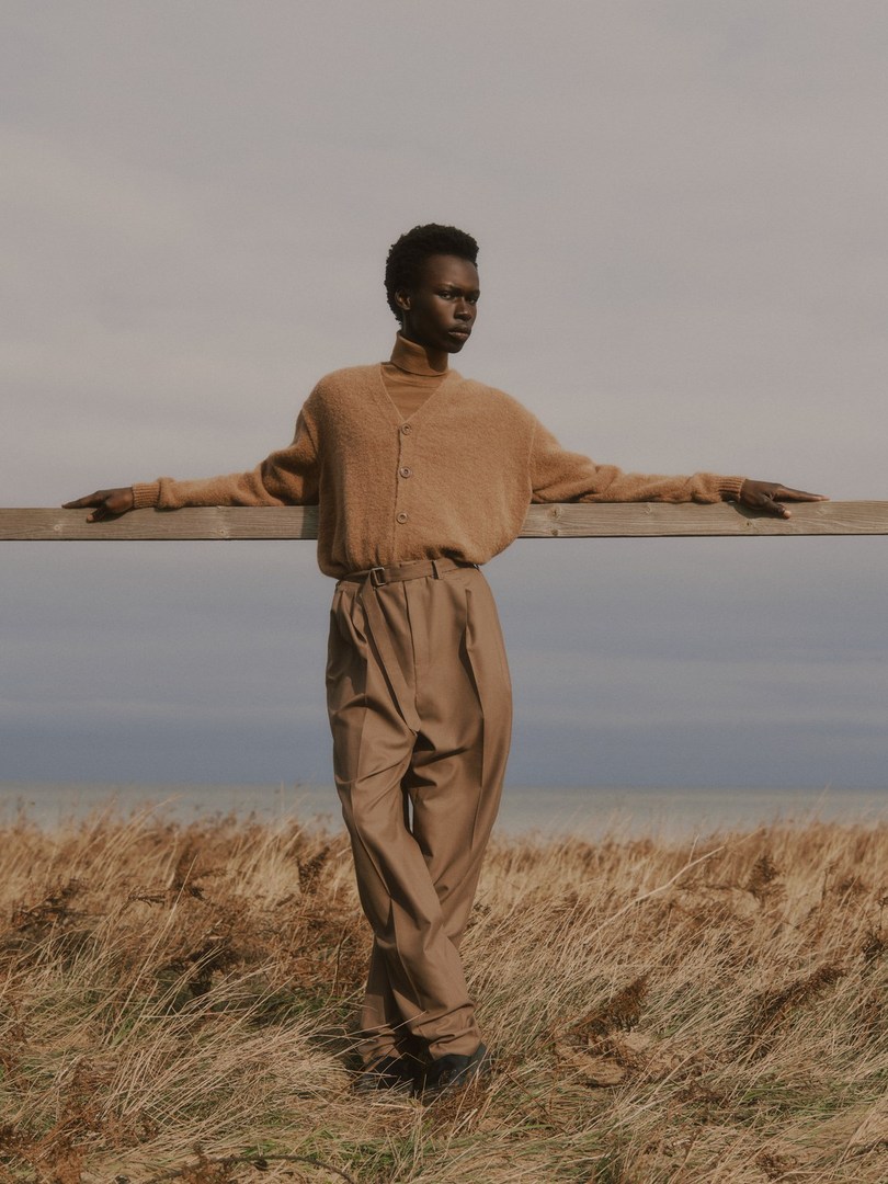 A dark-skinned model wears a brown cardigan and wide-legged pants in a field of grass, leaning on a wooden fence with the ocean in the background.