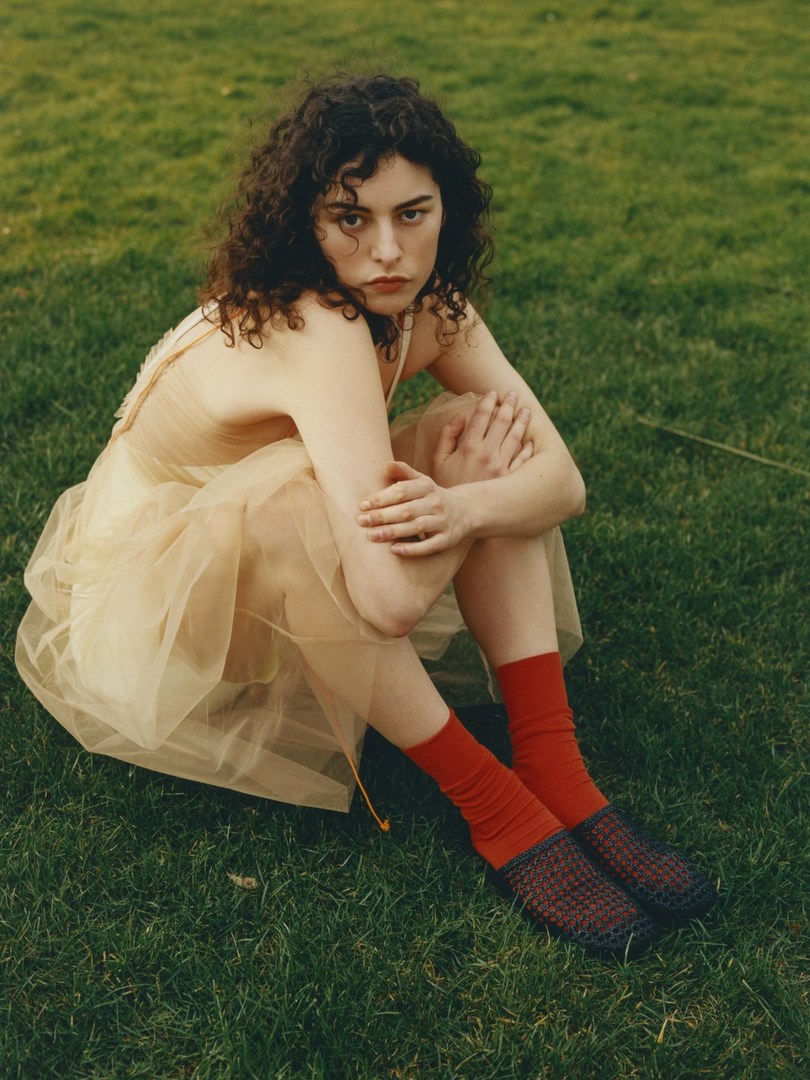 A young woman with curly hair sits cross-legged on green grass, wearing a light dress with red socks and dark, woven shoes in an outdoor scene.