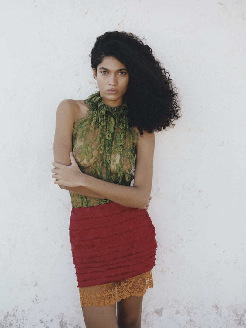 A woman with dark curly hair poses against a white wall, wearing a sheer green floral top and a layered red and orange skirt.