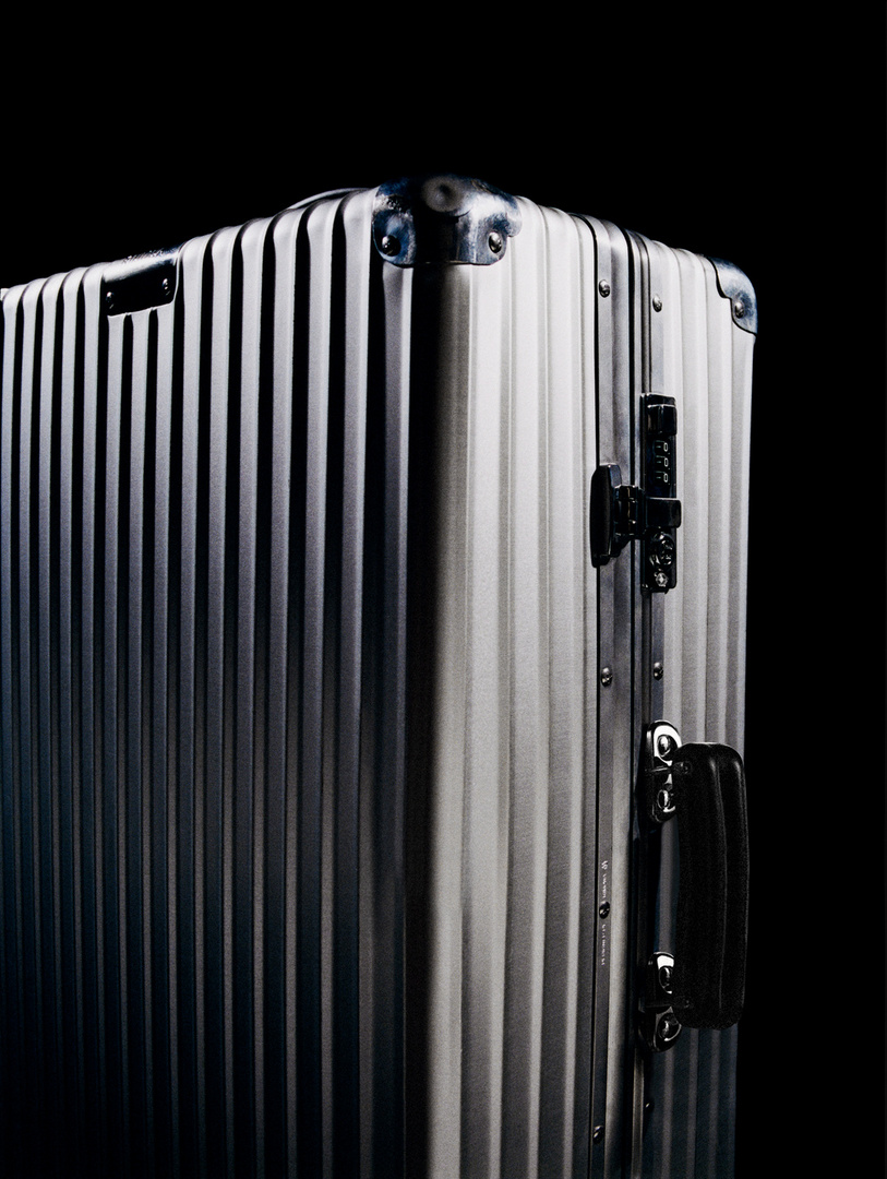 Angled close-up of shiny, ridged aluminum luggage with black handle and corner guards against a solid black background.