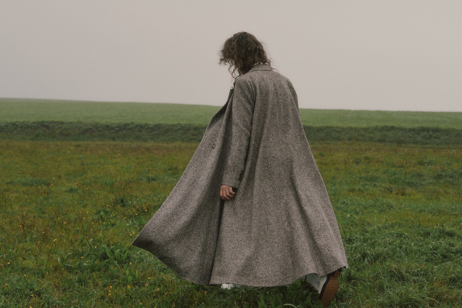 A woman walks across a green field wearing a long grey coat, her back to the camera, with fog in the background in a landscape shot.