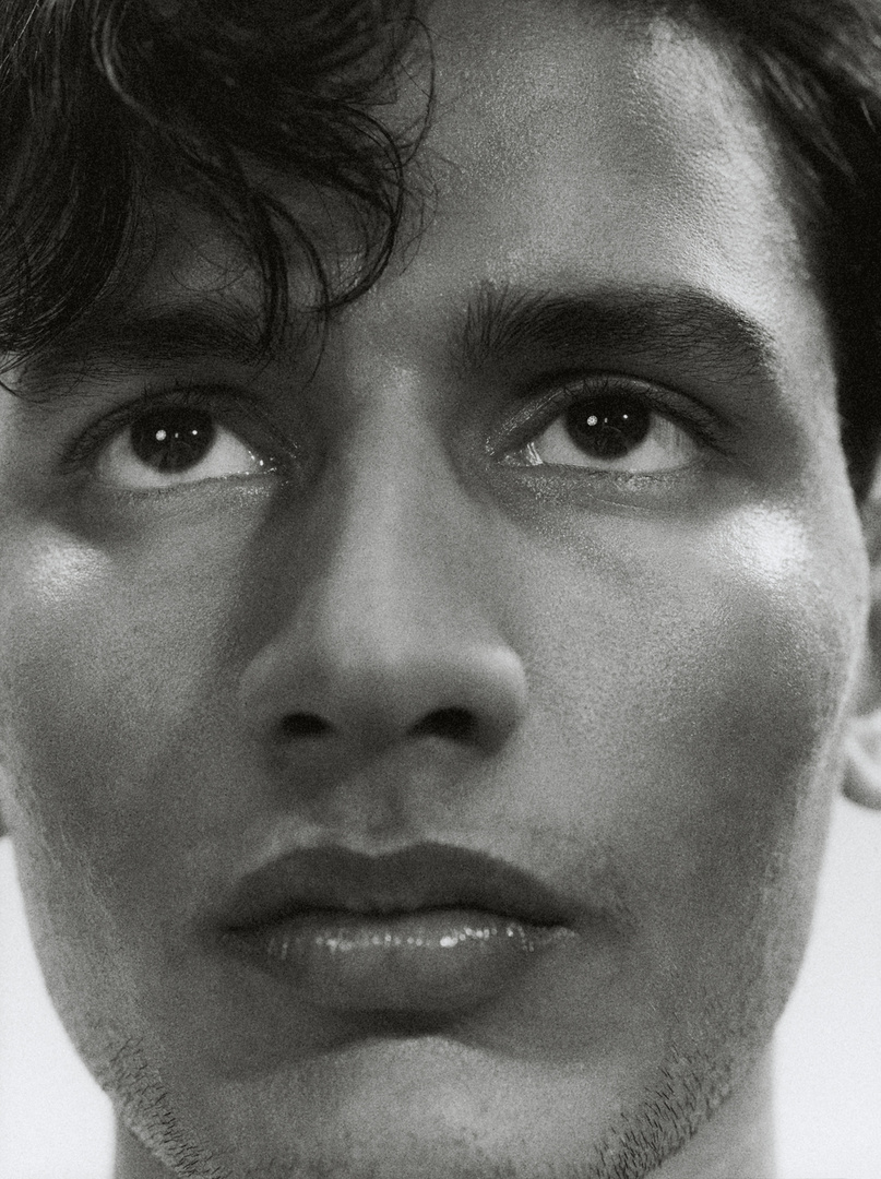 Close-up black and white portrait of a young adult male looking upwards with a neutral expression, highlighting his facial features and curly hair.