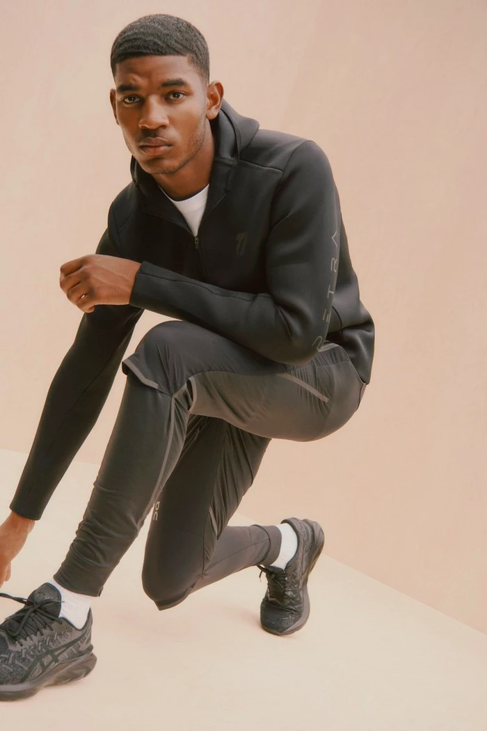 Black man crouching in black athletic hoodie and trousers against a warm neutral studio background.