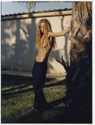 Long-haired model leaning against a palm tree trunk wearing dark trousers outdoors near a wall at sunset.