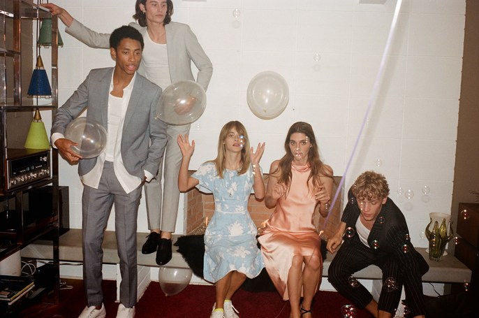 Group of five young adults, three standing and two seated, formally dressed with balloons and bubbles, indoor setting with neutral color palette.