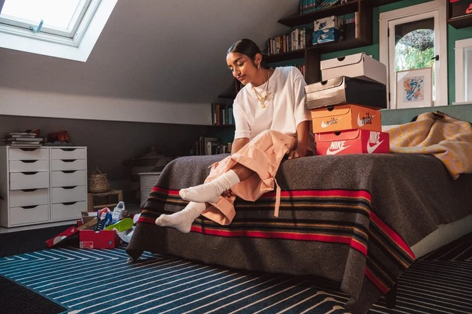 Young woman in white shirt and light orange pants sits on a bed with sneaker boxes in a room with a sloped ceiling and bookshelves.