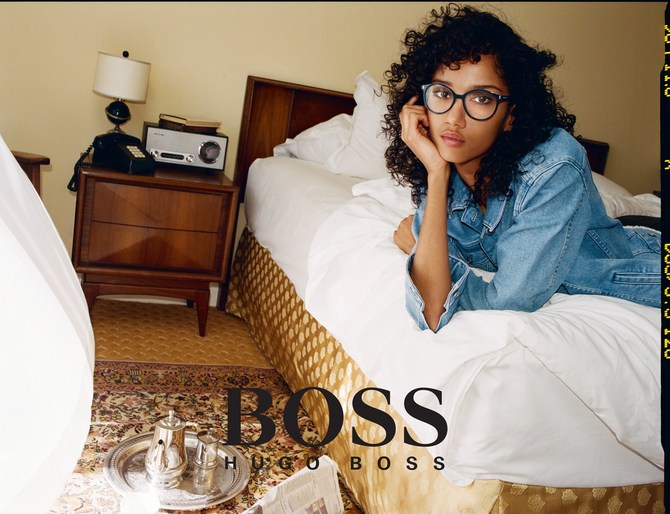 Young woman with curly hair poses in a denim jacket on a bed in a hotel room with a vintage aesthetic, promoting the BOSS brand.