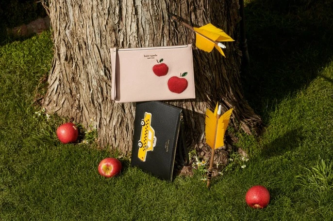 Two purses featuring apple and taxi designs sit beside a tree with scattered red apples and yellow arrows on green grass.