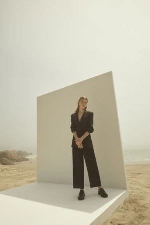 Model wearing a dark blazer and trousers standing on a white geometric platform on a sandy beach.
