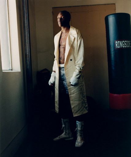 Man in boxing attire poses with boxing gloves and boots next to a black punching bag in a sparse locker room with natural light.