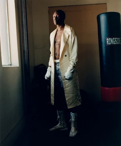 Man in boxing attire poses with boxing gloves and boots next to a black punching bag in a sparse locker room with natural light.