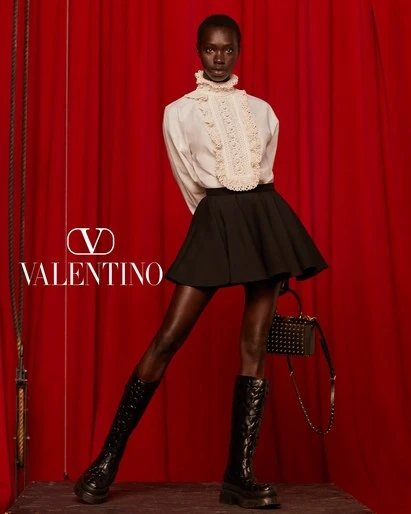 Valentino fashion ad: An African female model poses in a white blouse, black skirt, and black boots against a red curtain backdrop.